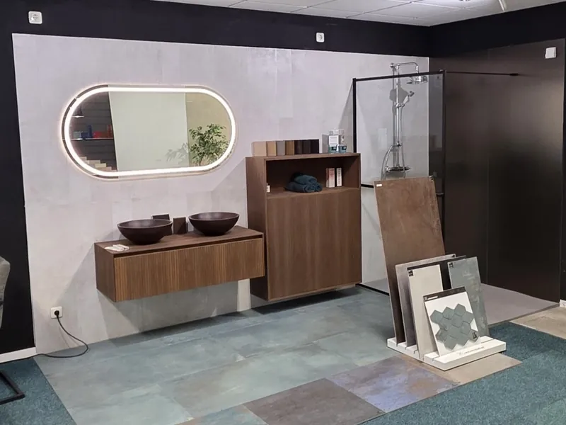 Wastafelmeubel van donker hout met twee waskommen, een verlichte ovale spiegel en een inloopdouche in de showroom van MIJS Tegels & Sanitair in Zwaag