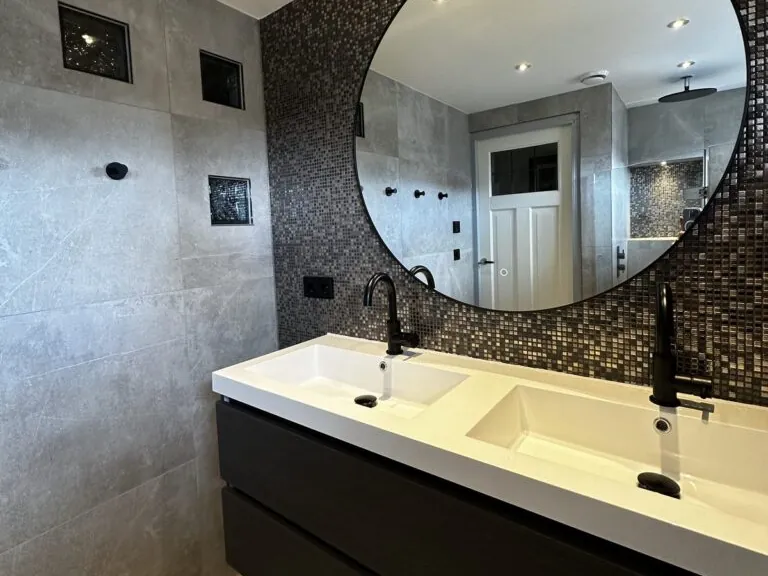 Badkamer met zwart mozaïek, ronde spiegel en matzwarte kranen, gerealiseerd door MIJS Tegels & Sanitair